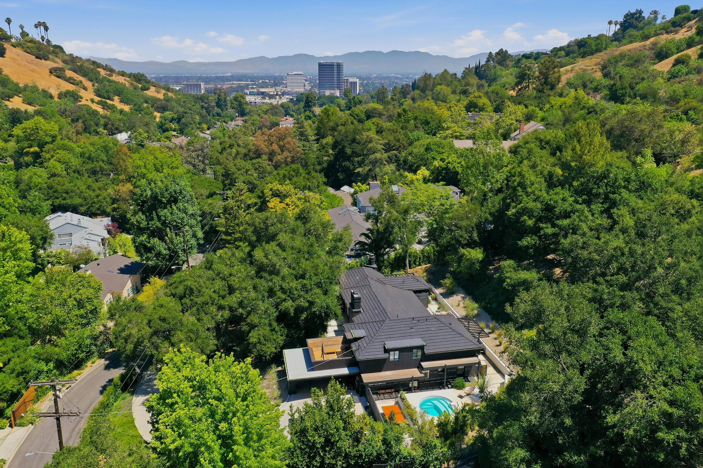 Residential drone view showing a hillside home, surrounding trees, and Los Angeles skyline